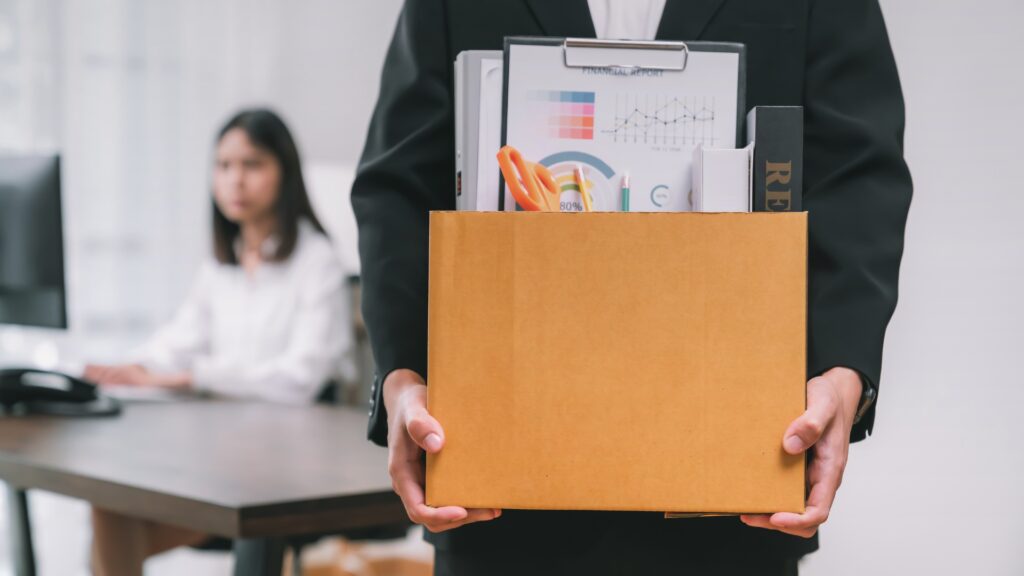 Hombre con caja de pertenencias laborales tras ser despedido, mientras una mujer observa desde un escritorio.